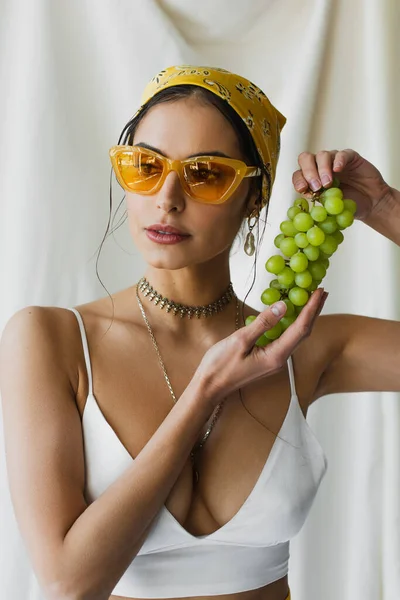 Trendy woman in yellow sunglasses, headscarf and crop top holding grapes on white — Stock Photo