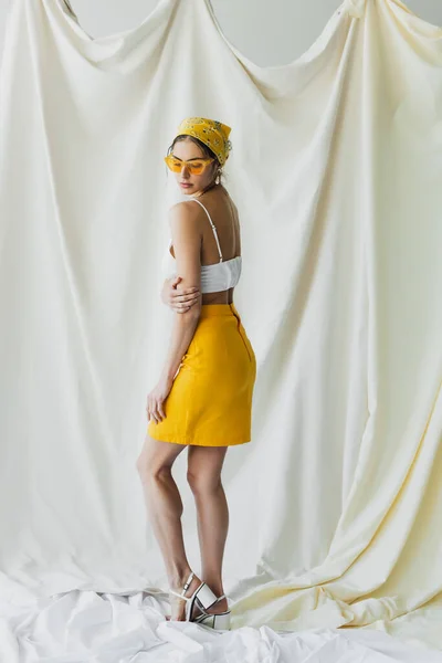 Longitud completa de la mujer bonita en gafas de sol amarillas y pañuelo en la cabeza posando en blanco - foto de stock