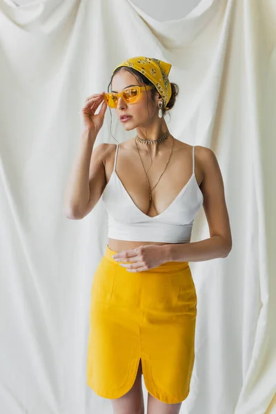 Mujer elegante ajustando gafas de sol y posando en blanco - foto de stock