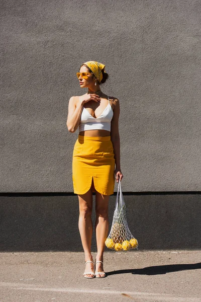 Piena lunghezza della donna elegante in occhiali da sole e velo giallo tenendo borsa stringa con frutta vicino muro di cemento — Foto stock