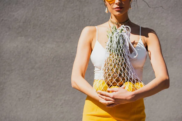 Vista ritagliata di donna elegante tenendo borsa stringa con frutta biologica vicino muro di cemento — Foto stock