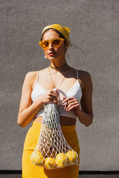 Stylish woman in yellow sunglasses and headscarf holding string bag with fresh fruits near concrete wall — Stock Photo