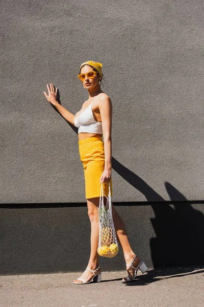 Pleine longueur de femme à la mode en lunettes de soleil et foulard jaune tenant un sac à ficelle avec des fruits près du mur en béton — Photo de stock