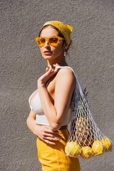 Mulher na moda em óculos de sol e lenço de cabeça amarelo segurando saco de corda com frutas perto da parede de concreto — Fotografia de Stock