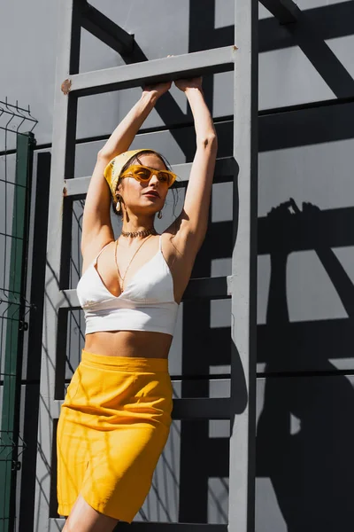 Jolie femme en lunettes de soleil et foulard jaune posant près de l'échelle et mur en béton — Photo de stock