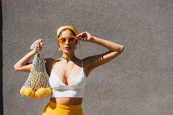 Stylish woman adjusting sunglasses and holding string bag with lemons and pineapple near concrete wall — Stock Photo