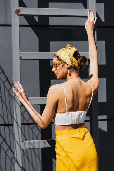 Femme élégante en lunettes de soleil et foulard jaune posant près de l'échelle et mur en béton — Photo de stock