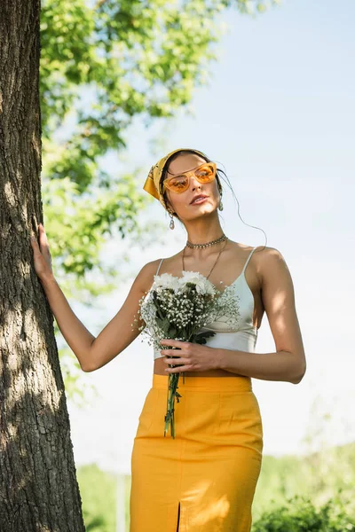 Femme élégante en lunettes de soleil et foulard jaune tenant des fleurs et debout près du tronc d'arbre — Photo de stock