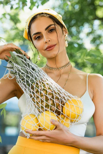 Mulher na moda em lenço de cabeça amarelo segurando saco de corda com frutas — Fotografia de Stock
