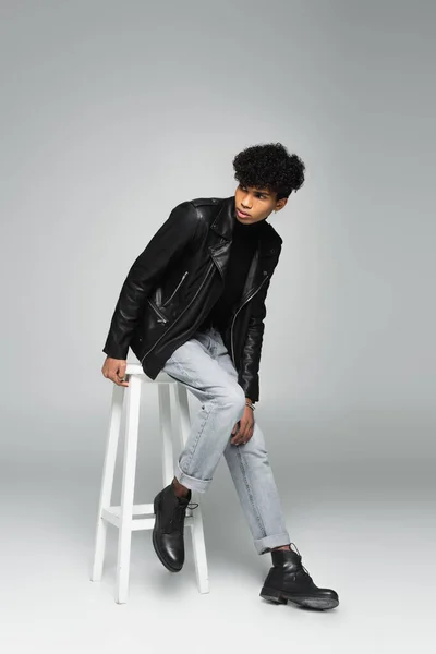 Trendy african american man in jeans and leather jacket looking away on high stool on grey background — Foto stock