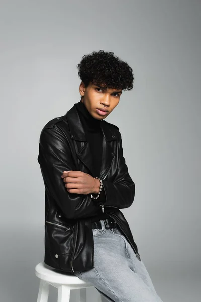 Trendy african american man in black leather jacket looking at camera while sitting isolated on grey — Fotografia de Stock