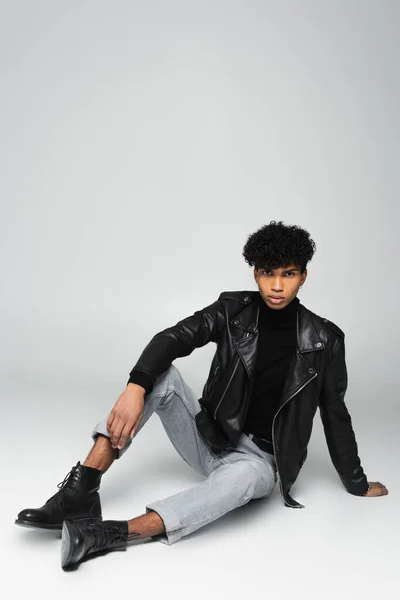 Молодой африканский американец в черной кожаной куртке и джинсах сидит на сером фоне — стоковое фото