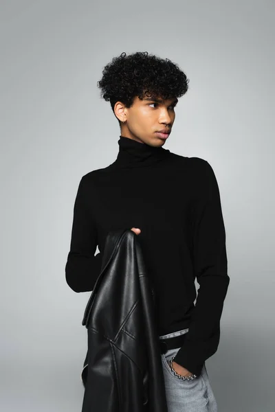 Young african american man in black turtleneck holding leather jacket isolated on grey - foto de stock