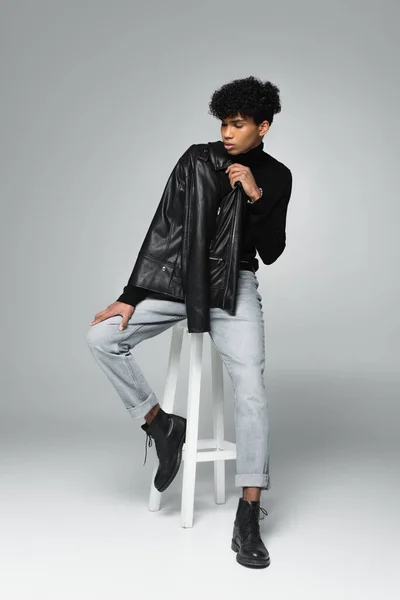 Young african american man in trendy clothes posing on high stool on grey background — Fotografia de Stock