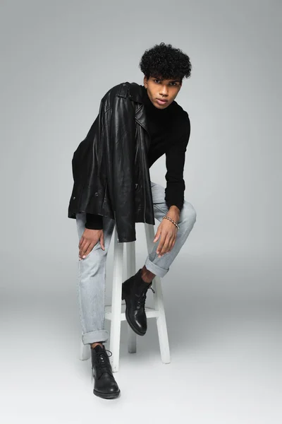 Young african american man with black leather jacket on shoulder posing on high stool on grey background — Stock Photo