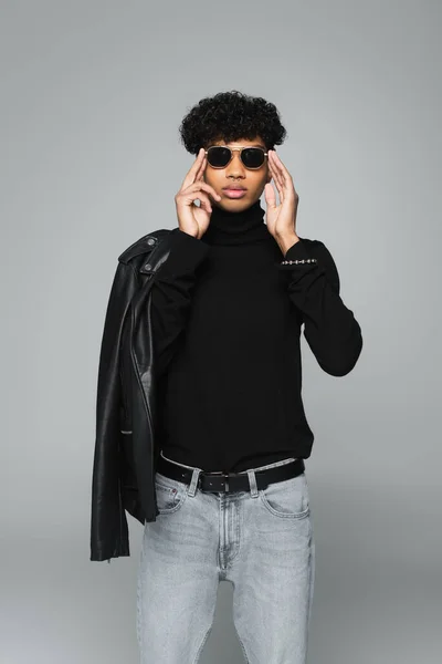 African american man in black turtleneck and jeans adjusting dark sunglasses isolated on grey — Foto stock