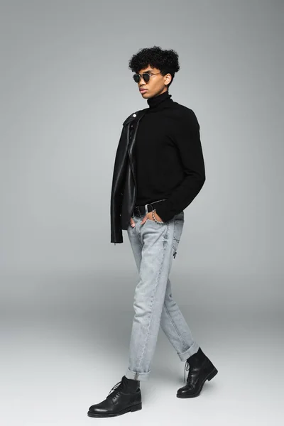 Trendiger afrikanisch-amerikanischer Mann in Jeans und schwarzem Rollkragen, der mit der Hand in der Tasche auf grauem Hintergrund geht — Stockfoto