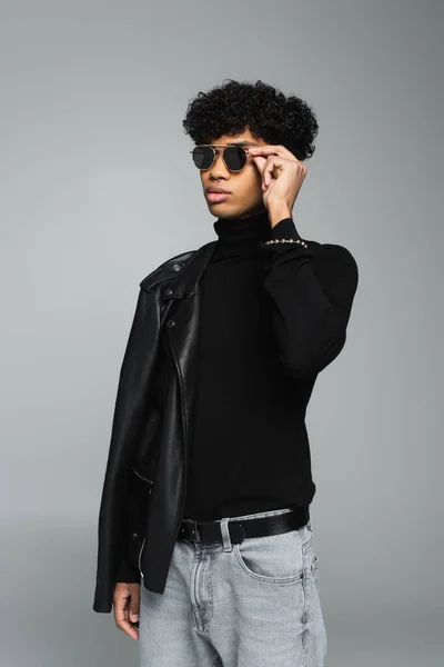 African american man in black turtleneck and leather jacket on shoulder adjusting dark eyeglasses isolated on grey — Photo de stock