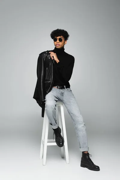 Young african american man in stylish clothes and black sunglasses sitting on high stool on grey background - foto de stock