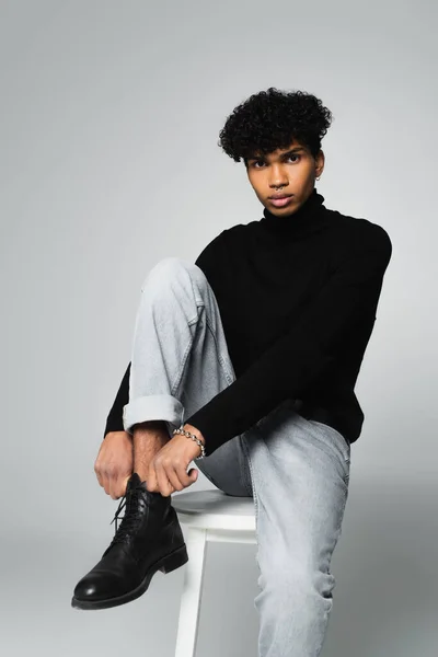 Trendy african american man in black turtleneck and jeans sitting on white stool and looking at camera on grey background — Photo de stock