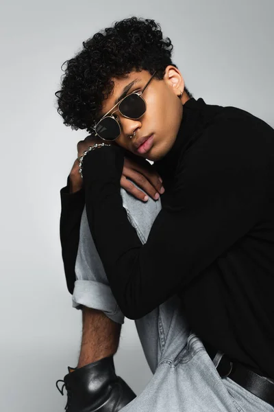 Young african american man in black turtleneck and dark sunglasses sitting isolated on grey - foto de stock