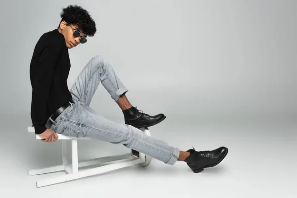 Trendy african american man in black turtleneck, jeans and dark sunglasses sitting on overturned stool on grey — Foto stock