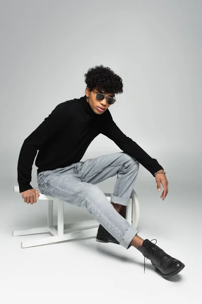 African american man in stylish clothes and black sunglasses posing on overturned stool on grey background — Photo de stock