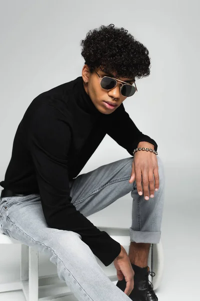 African american man in jeans, black turtleneck and dark sunglasses sitting on overturned stool on grey — Photo de stock