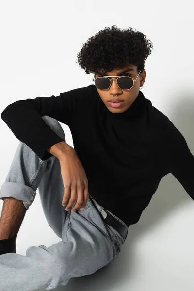 African american man in dark sunglasses and black turtleneck looking at camera while sitting on white — Fotografia de Stock