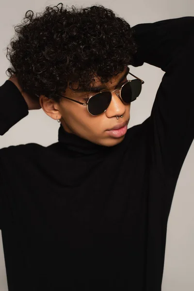 Pierced african american man in black turtleneck and dark sunglasses posing with hands behind head isolated on grey - foto de stock