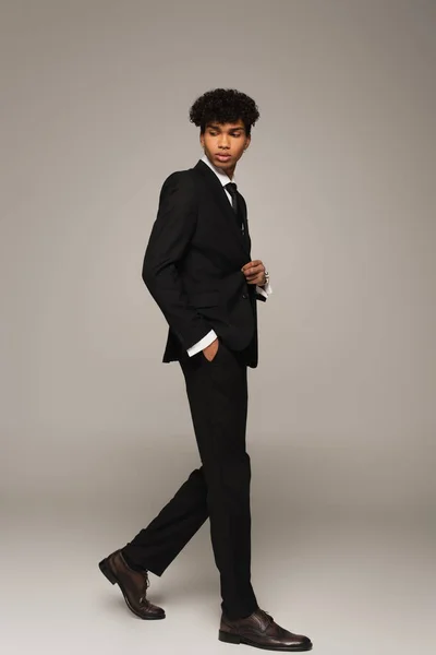 Young african american man in elegant suit and oxford shoes walking with hand in pocket on grey background — Fotografia de Stock