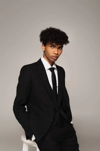 Elegant african american man with hands in pockets looking at camera while sitting isolated on grey — Stock Photo