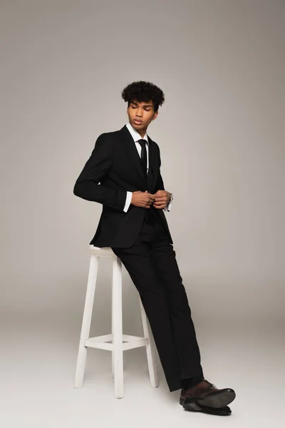 African american man in suite buttoning blazer while sitting on grey background - foto de stock