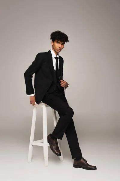 Full length view of african american man in elegant formal wear sitting on high stool on grey background — Photo de stock