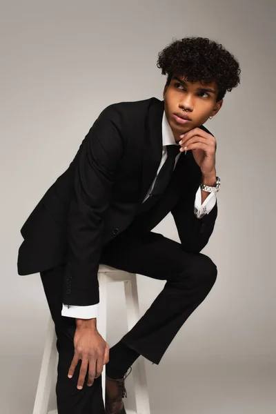 Afrikanisch-amerikanischer Mann im schwarzen eleganten Anzug schaut weg, während er isoliert auf grau sitzt — Stockfoto