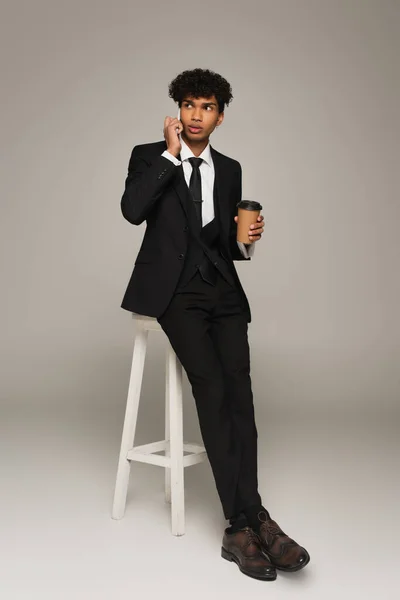 Afrikanisch-amerikanischer Geschäftsmann sitzt auf einem hohen Hocker mit Coffee to go und telefoniert auf grauem Hintergrund — Stockfoto