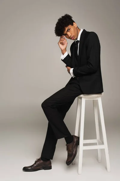 Young african american man in black suit sitting on white stool and looking at camera on grey - foto de stock