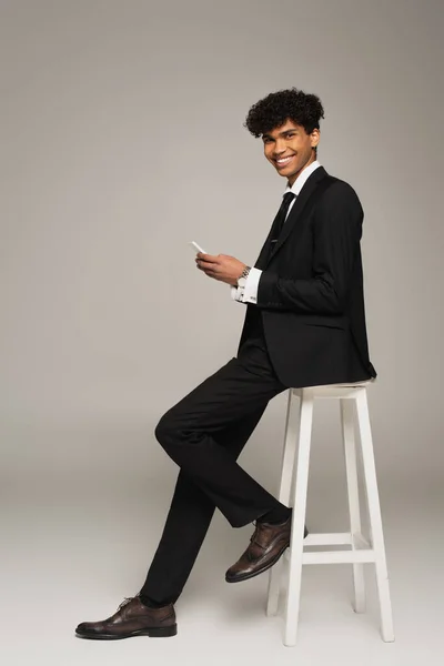 Елегантний афроамериканський чоловік з мобільним телефоном посміхається на камеру, сидячи на сірому фоні — стокове фото