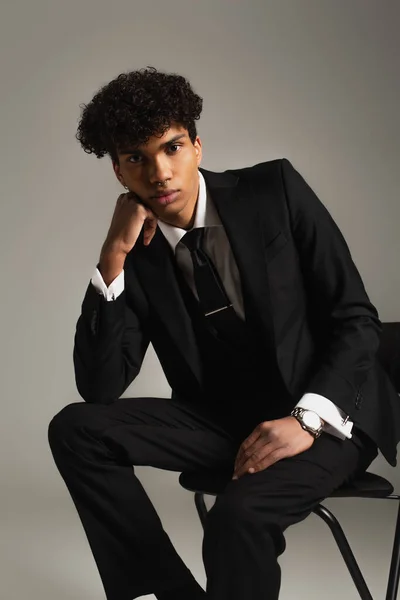 African american man in black elegant suit looking at camera while sitting on chair isolated on grey - foto de stock