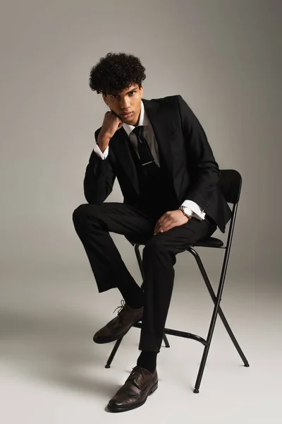 Trendiger afrikanisch-amerikanischer Mann im eleganten Anzug sitzt auf einem Stuhl mit händennahem Gesicht auf grauem Hintergrund — Stockfoto