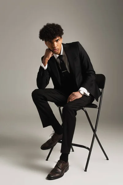 Trendy african american man in elegant suit sitting on chair with hand near face on grey background - foto de stock