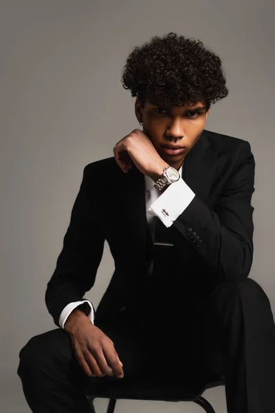 Stylish african american man in black suit looking at camera while sitting isolated on grey — Stock Photo