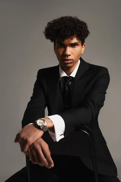 Young african american businessman in black elegant suit sitting on chair and looking at camera isolated on grey — Fotografia de Stock