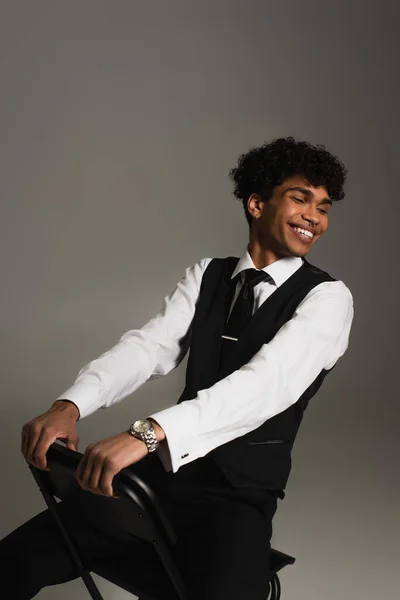 Elegant african american man smiling with closed eyes while sitting isolated on grey — Fotografia de Stock