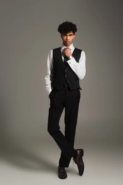 Elegant african american man adjusting black tie while posing with hand in pocket on grey background - foto de stock