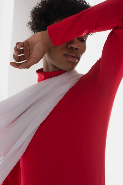 Young african american man in red turtleneck covering face with arm while posing near white drapery — Foto stock