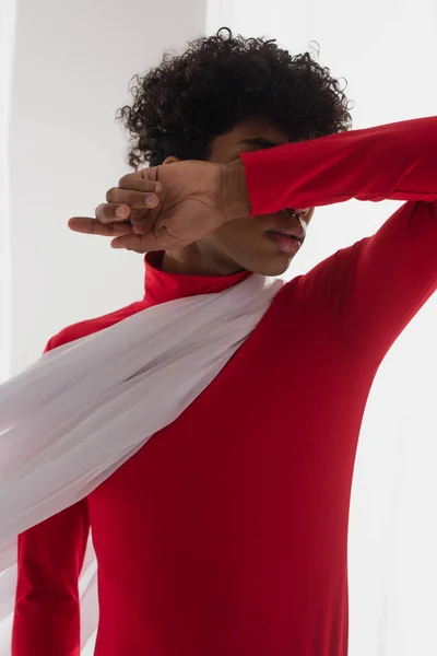 African american man in red turtleneck obscuring face with arm while posing in white chiffon cloth — Stock Photo