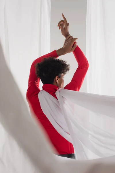 Back view of african american man with trendy hairstyle posing near white cloth with raised hands — Fotografia de Stock