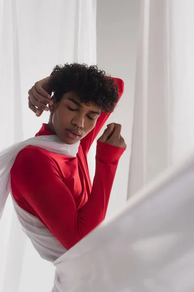 African american man in red turtleneck posing with closed eyes near white tulle drapery — Fotografia de Stock