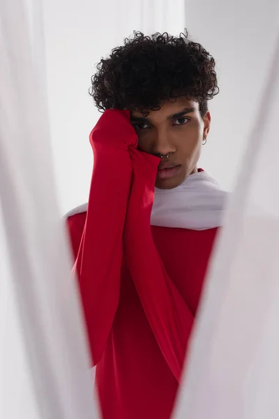 Young african american man in red turtleneck posing near white chiffon cloth with hands near face — Foto stock